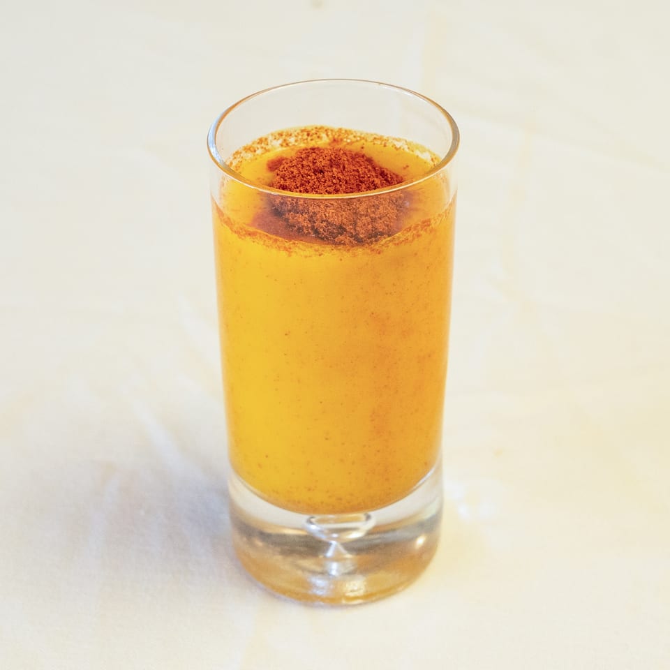 Yellow-orange drink in a tall glass, topped with reddish-brown powder, on a light background.