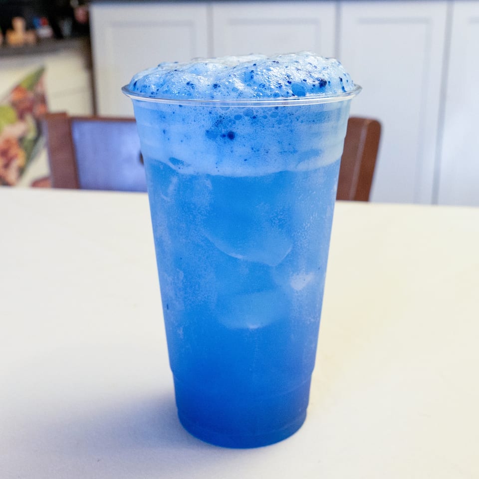Vibrant blue foamy drink with ice in a plastic cup on a white table.