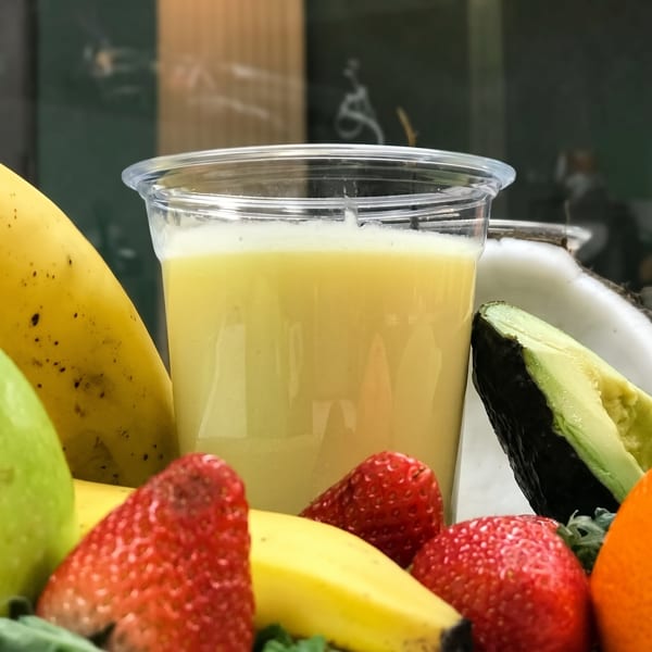Yellow smoothie in a clear cup surrounded by fresh bananas, strawberries, avocado, and coconut.
